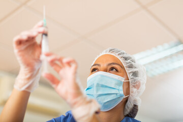 Hispanic female doctor in face mask preparing syringe for vaccination in hospital
