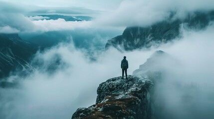 A lone figure stands on a rocky outcrop, overlooking a dramatic landscape shrouded in mist and clouds. The person is seen from the back, dressed in dark outdoor clothing with a backpack, suggesting an