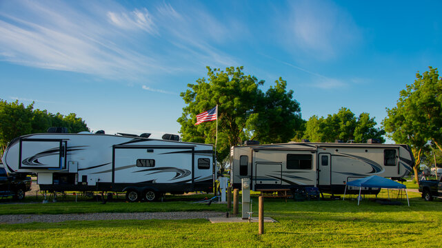 Fifth wheel trailers parked back to back at campsite on grass with trees and blue cloudy skies
