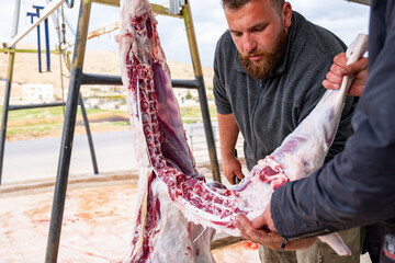 A bearded man is skillfully butchering a sheep that is hanging on a hook, carefully preparing it for consumption