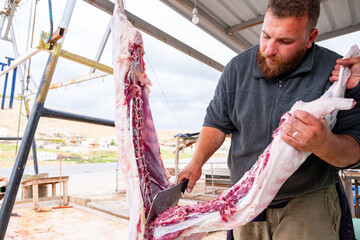 The butcher is cutting meat outdoors, holding his knife, and smiling with a bearded face