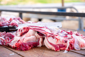 In the springtime, a bearded butcher is cutting meat at a wooden table outdoors, with sheep in the...
