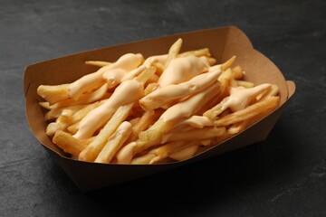 Tasty potato fries and cheese sauce in paper container on black table, closeup