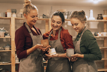 A company of three female friends in a ceramic workshop shows each other homemade mugs.
