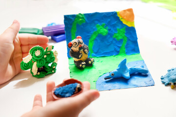 Naklejka premium Child smearing colorful plasticine on cardboard and creating fairy tale card with cartoon animals, panda, birds, shark. By spreading, modelling and adding texture to plasticine child creating scene