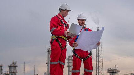 Engineer wear uniform and helmet stand workplace hand holding tablet computer, survey inspection team work see detail blue print plant site to work with oil refinery  background. - 800795715