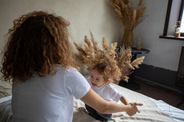 happy ethnic curly mom playing with laughing little baby daughter in bed, playful little girl merrily looking at mother