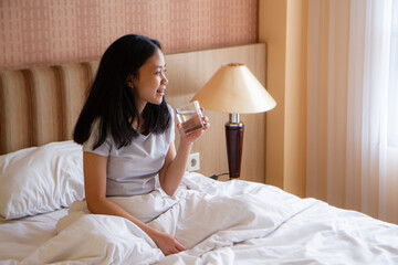 beautiful happy asian girl holding drinking water from glass while sitting on bed in the morning.