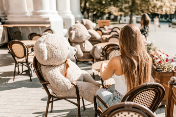A woman sits cafe with a teddy bear next to her. The scene is set in a city with several chairs and tables around her. The woman is enjoying her time at the outdoor cafe.
