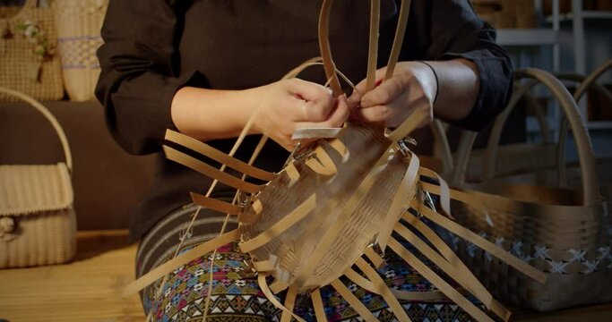 Slow motion scene while a beautiful Asian female artisan who is a local villager weaving folk basket made from leftover paper band to sell as extra income in the basketry shop shop.