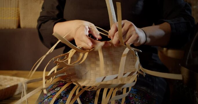 Slow motion scene while a beautiful Asian female artisan who is a local villager weaving folk basket made from leftover paper band to sell as extra income in the basketry shop shop.