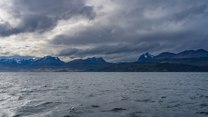 The picturesque snow-capped Andean Martial mountain range is visible from the side of the Beagle Canal. The city of Ushuaia is at the foot of the mountains on the ocean. Seabirds fly over the water