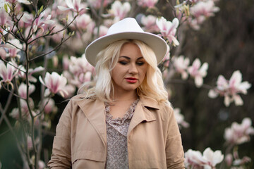 Magnolia flowers woman. A blonde woman wearing a white hat stands in front of a tree full of pink flowers. She is wearing a brown jacket and a necklace.