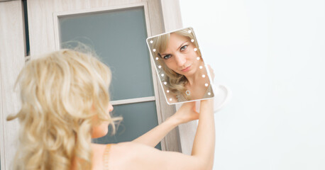 A woman looks at herself in the mirror. She is wearing a white shirt and her hair is styled so that...