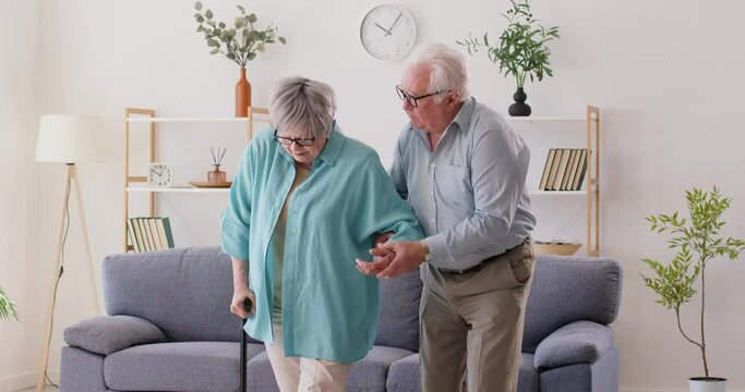 Tender support in every step. Devoted elderly man helps his mature wife, who walks with walking cane, across room. Caucasian man supports sick woman with cane. Retirement health and illness concept.