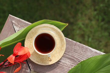 tea cup, tropical green canna leaves and red flower on wooden balcony railing. tea time, morning, relaxation. top view - obrazy, fototapety, plakaty