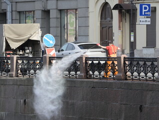 A cleaner in an orange vest washes the embankment with water from a hose, Moika River Embankment,...