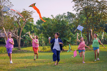 Happy Asian Thai group of boys and girls wear traditional colorful farmer outfits and run with flying kites on a green field.