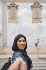 Vertical photo of female tourist standing holding hands with scarf and backpack smiling looking at camera near church with famous Portuguese blue ceramic tiles. Honeymoon.