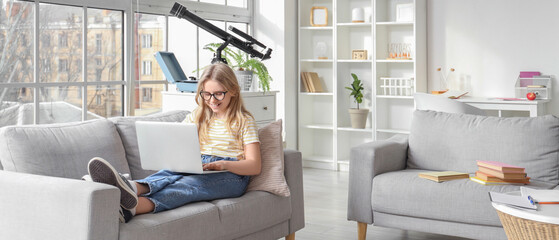 Cute little girl in eyeglasses using laptop on sofa at home