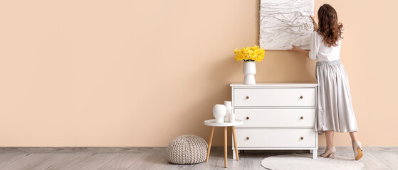 Young pretty woman hanging picture on beige wall near chest of drawers in room