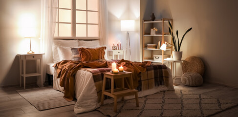 Interior of bedroom with brown checkered blankets on bed, burning candles and glowing lamps late in...