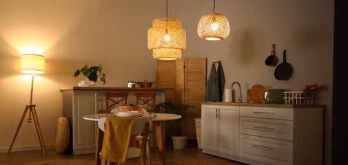 Interior of modern kitchen with glowing lamps, dining table and white counters at evening