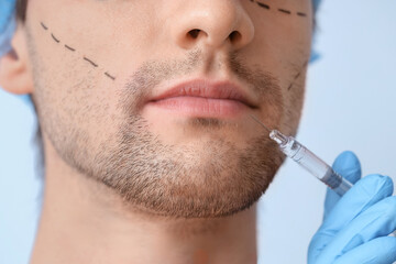 Young man with marked face receiving lip injection on blue background, closeup. Plastic surgery...