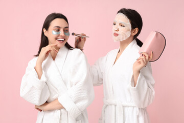Female friends in bathrobes making cosmetic procedures to each other on pink background