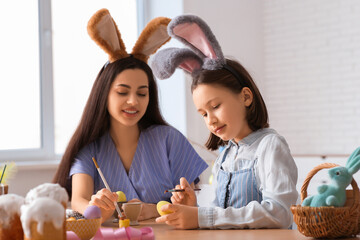 Little girl and her mother in bunny ears painting Easter eggs at kitchen