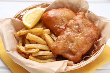 Tasty fish, chips and lemon in wicker bowl on white wooden table, closeup