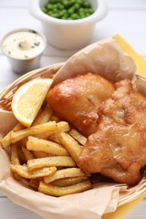 Tasty fish, chips, sauce and lemon in wicker bowl on table, closeup