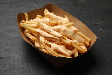 Tasty potato fries and cheese sauce in paper container on black table, closeup