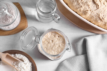 Sourdough starter in glass jar, flour, dough and water on light table, flat lay