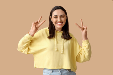 Smiling young woman showing victory gesture on brown background