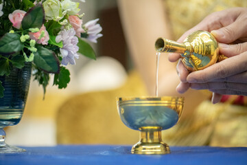 The groom is pouring water from a golden vessel into a golden bowl during a Thai wedding ceremony