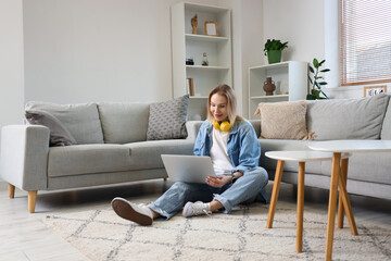 Fototapeta premium Young woman in headphones using laptop on floor in light living room