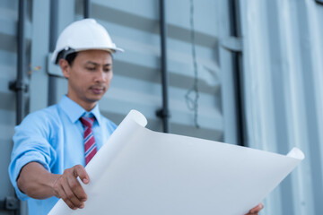 An engineer wearing a hard hat and holding a blueprint is looking at the construction site.