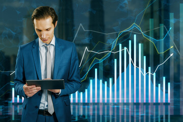 Attractive young businessman using tablet with growing forex chart on blurry city background....