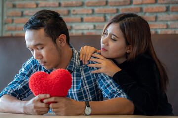 A man and a woman are sitting at a table. The man is holding a red heart-shaped pillow and looking...