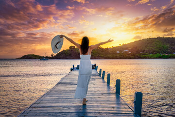 A beautiful woman in a white dress and with hat enjoys the tropical sunset in the Caribbean Sea,...