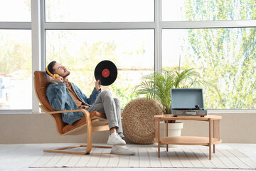 Young bearded man in headphones with vinyl disk listening to music on armchair at home