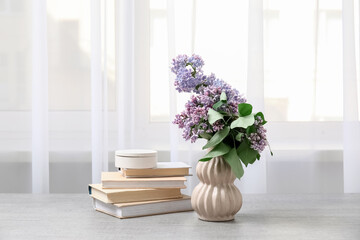 Books and vase with lilacs branches on table near window in room