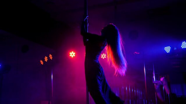 Silhouette of a girl with long flowing hair dancing on a pole in a dark smoke-filled strip club. Slow motion. Pole dance.