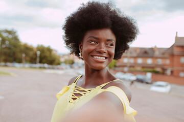 Portrait of a young smiling afro woman taking selfie at city park. Wears yellow top and holds...