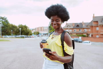 Young cheerful african female student wears yellow top, afro hairstyle and backpack, drinks coffee...