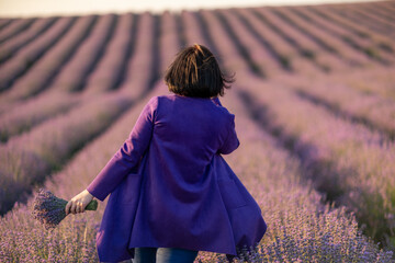A woman in a purple coat is walking through a field of lavender. The scene is serene and peaceful, with the purple flowers creating a calming atmosphere. The woman is enjoying her time in the field.