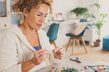 Young mature youthful woman enjoying at home with indoor leisure hobby activity making jewels and necklaces with colorful beads and watching a tablet device to follow tutorial instructions online.