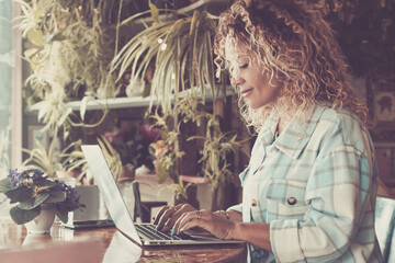 Side view of young woman sitting at the desk and writing on laptop. One businesswoman in modern smart working. Garden plants store shop. Female people using computer indoor to work and enjoy business