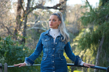 Pretty young blonde woman with a ponytail in her hair wearing a denim dress. Trees and plants in...